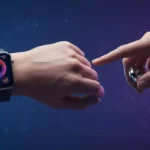 Um smartwatch cinza-escuro no pulso esquerdo de uma pessoa, exibindo um mostrador colorido com gráficos de saúde. A mão direita de outra pessoa aponta com o dedo indicador para a tela do smartwatch, enquanto usa um anel inteligente prateado com três LEDs azuis no dedo médio. O fundo é um degradê de azul e roxo com um padrão hexagonal sutil.